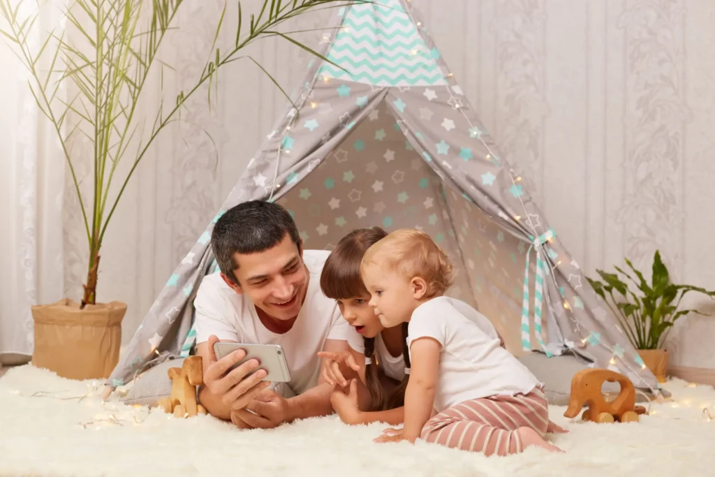 horizontal shot smiling man white t shirt laying floor with children holding smart phone hands kids looking mobile phone display with astonished expressions min scaled