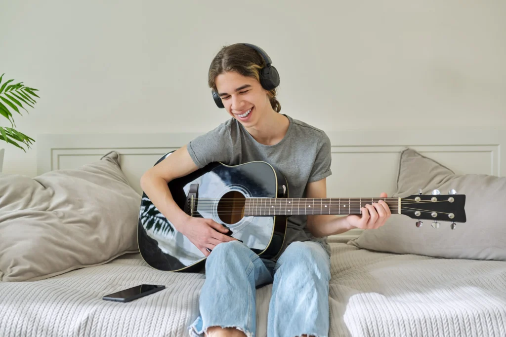 male teenager headphones sitting home with acoustic guitar smartphone min scaled